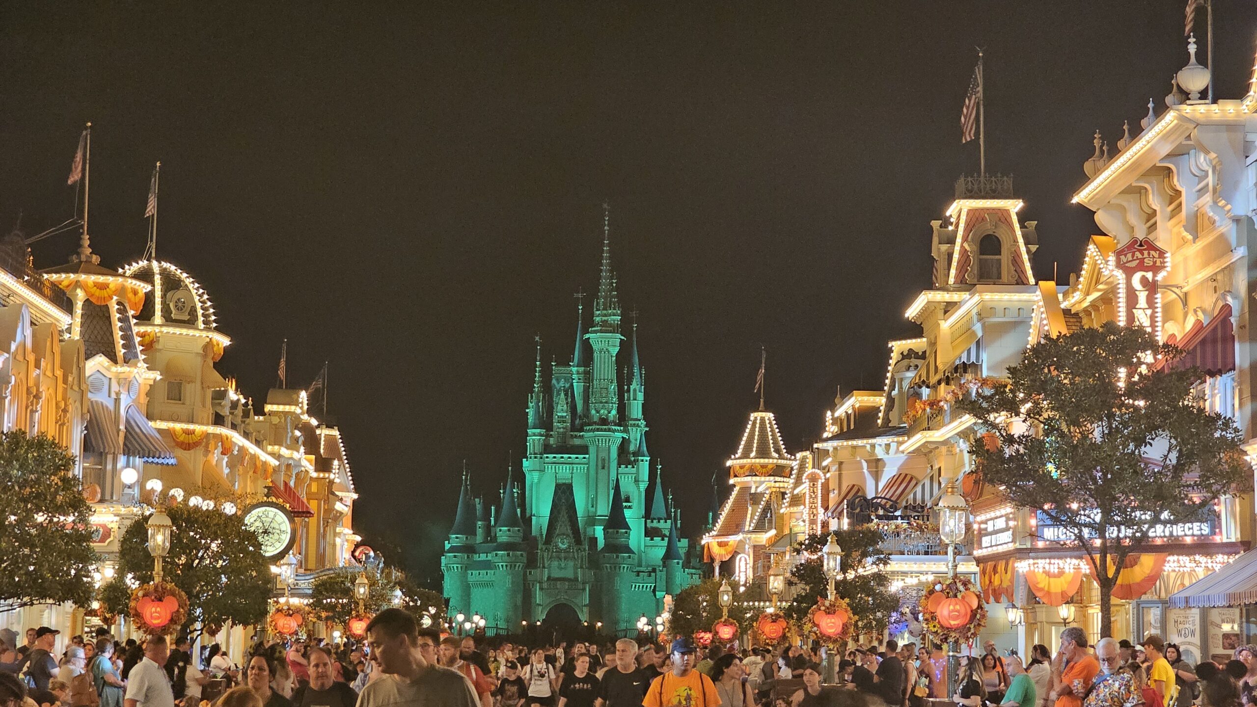 The buildings of Main Street U.S.A. at night are lit up along the sides of the photo with Cinderella Castle in the center. The castle is lit up green and orange and yellow banners and Mickey-shaped pumpkin lights decorate Main Street for fall. Crowds of party-goers are also seen on Main Street.