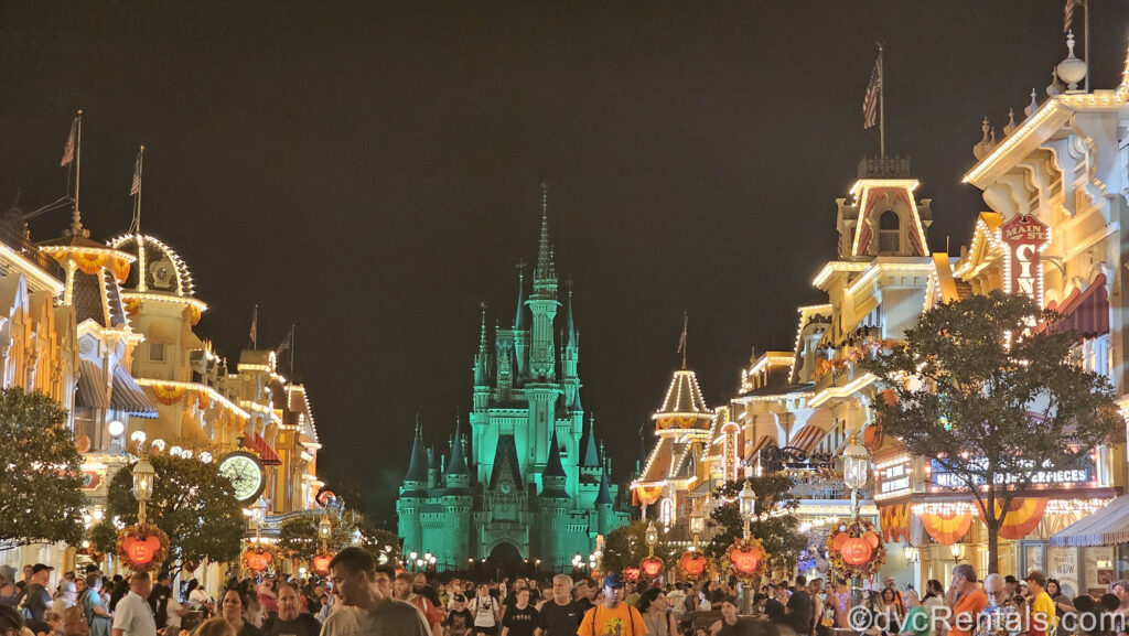 The buildings of Main Street U.S.A. at night are lit up along the sides of the photo with Cinderella Castle in the center. The castle is lit up green and orange and yellow banners and Mickey-shaped pumpkin lights decorate Main Street for fall. Crowds of party-goers are also seen on Main Street.