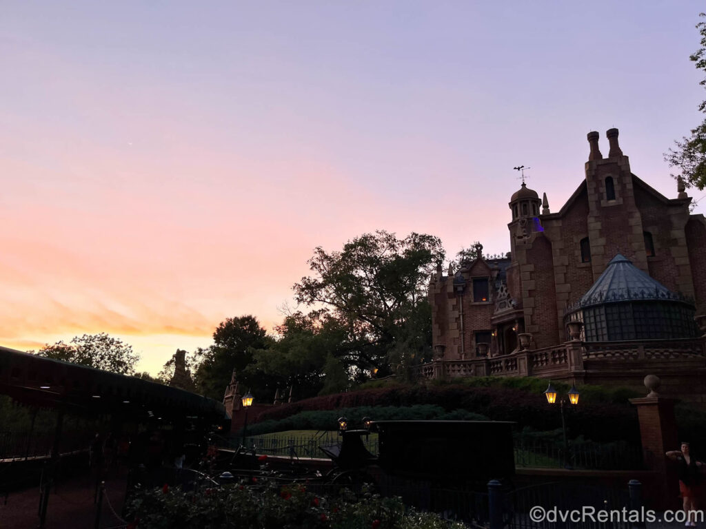 The red brick and beige stone building of the Haunted Mansion is seen under a pink and orange sunset. In front of the mansion there is green foliage made to look overgrown, as well as red roses in the garden and a black old-fashioned horse-drawn carriage.