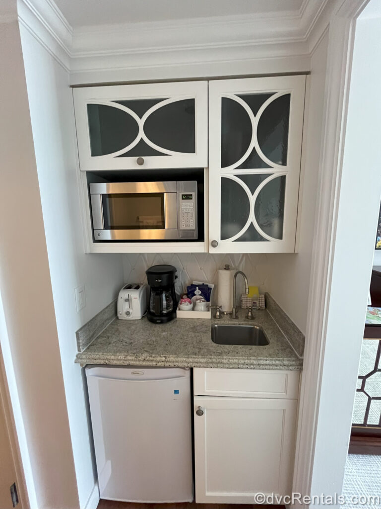 A kitchenette in a Deluxe Studio at the Villas at Disney’s Grand Floridian Resort & Spa. The speckled grey countertop holds a white toaster, black coffeemaker, cream and sugar and coffee packets, paper towel, sponge, and dishtowel next to the mini sink. Below the counter is a white cabinet and mini fridge. Above the counter are white cabinets with glass cut-outs and a stainless-steel microwave.