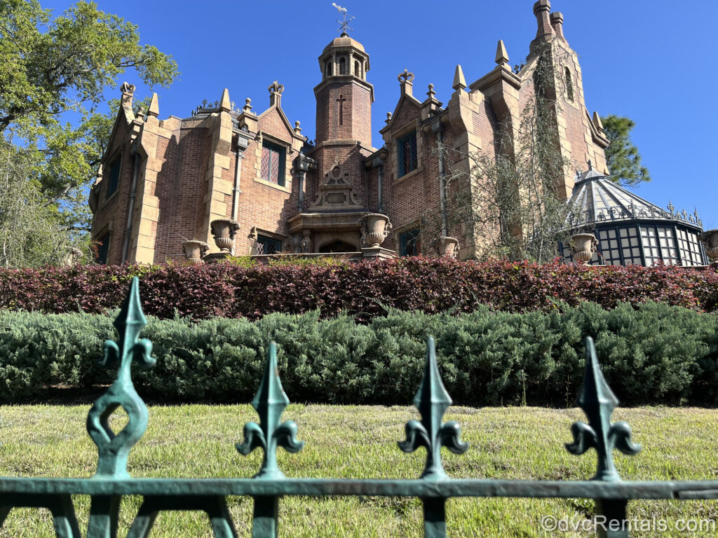 The red brick and beige stone building of the Haunted Mansion in daylight. In front of the mansion there is burgundy and green foliage, seen behind the green filigree spikes of the iron fencing in front of the attraction.