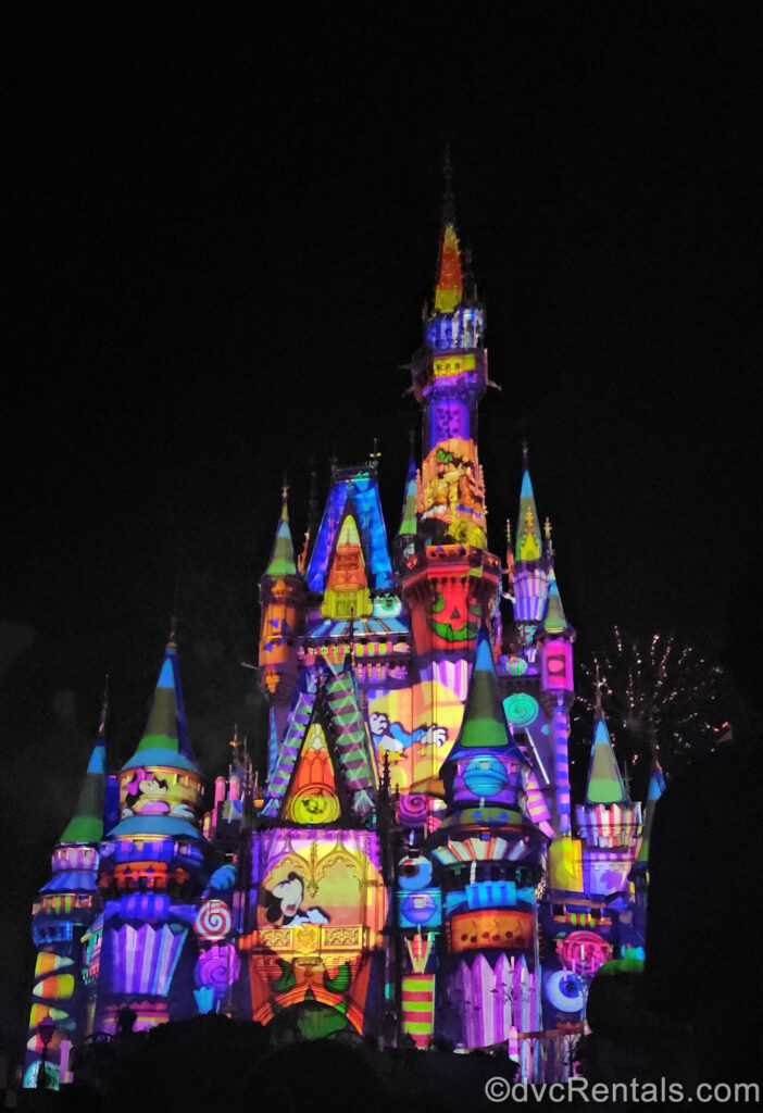 Colorful Halloween and candy-themed projections featuring Mickey Mouse, Minnie Mouse, Donald Duck, and Goofy light up Cinderella Castle, with fireworks sparkles in the background.