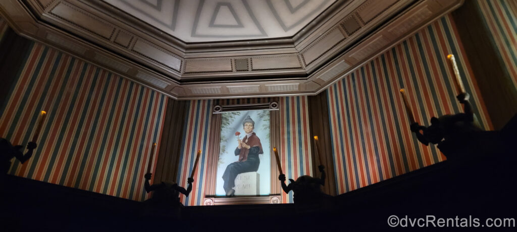 The Stretching Room of the Haunted Mansion. There is blue and red striped wallpaper and shadowy gargoyles holding flickering taper candles. On the wall there is a portrait of a grey-haired woman holding a rose and sitting on a tombstone