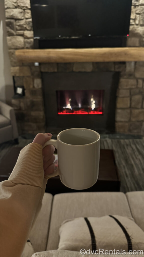 A hand holds a white coffee cup in front of the stone fireplace in the living room area of a Cascade Cabin at the Copper Creek Villas & Cabins at Disney’s Wilderness Lodge. The beige sofa can be seen below the cup, and a small fire flickers in the fireplace.