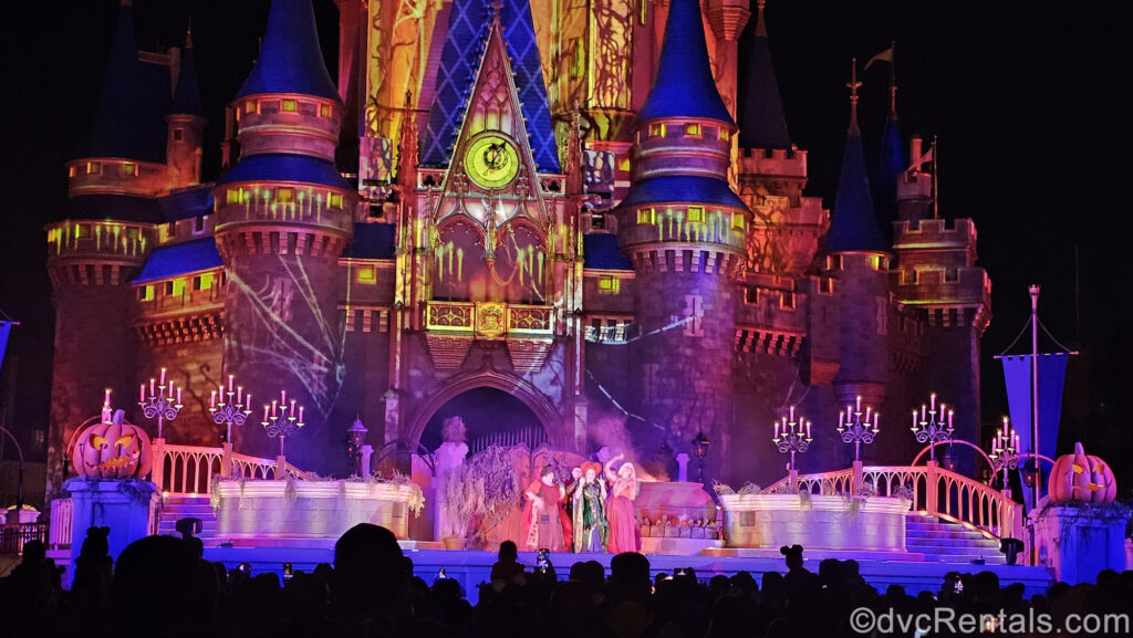 The Sanderson Sisters perform on the stage at Cinderella Castle at Magic Kingdom. The castle is lit up with yellow and pink projection effects with big cobwebs, and there are several large candelabras and big jack o’ lanterns decorating the stage.