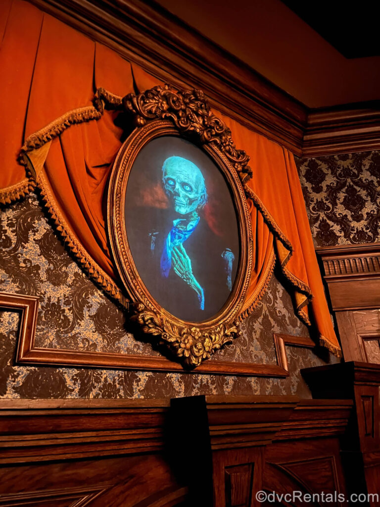 A portrait of a man transforming into a skeleton hangs in a bronze frame. There is black and gold damask-style wallpaper, a crimson curtain, and carved wood paneling that also decorate the wall the portrait hangs on.