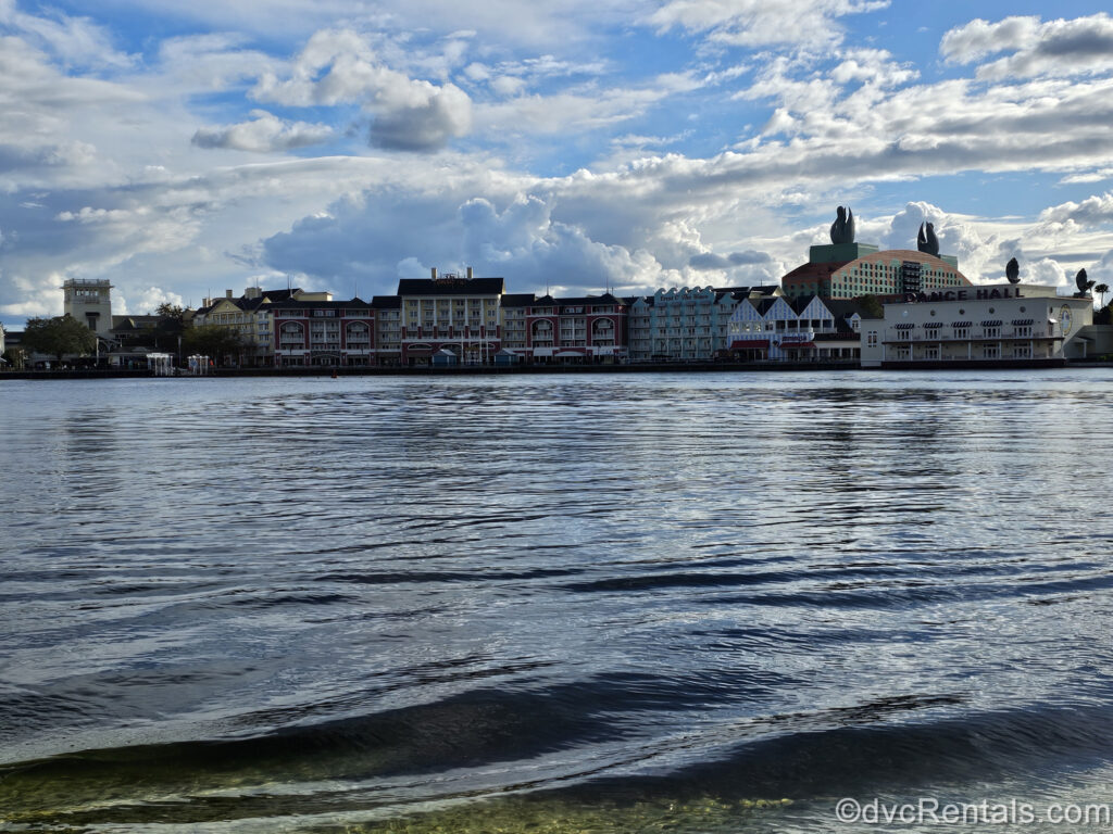 The colorful buildings of Disney’s BoardWalk Villas seen from across the waters of Cresent Lake under a sky full of fluffy white clouds and sunshine.