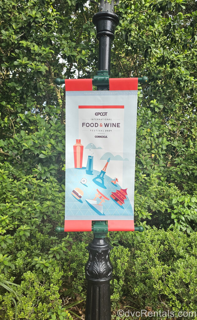 A red, white, and blue banner reading “Epcot International Food & Wine Festival 2024 presented by Corksicle” hangs on a black lamppost in front of green foliage.