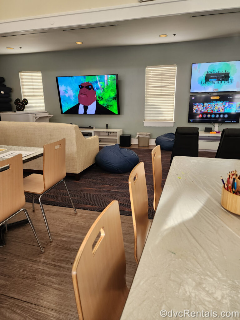 The interior of the Community Hall at Disney’s BoardWalk Villas. There is a movie playing on TV in front of a beige couch, video game screens, and a wooden table with coloring supplies.