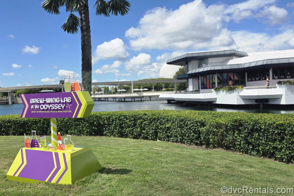 The lime green and bright purple sign for the Brew-Wing Lab at the Odyssey sits on green grass at EPCOT. The white Odyssey building is visible in the background along the waters at the edge of World Showcase Lagoon.