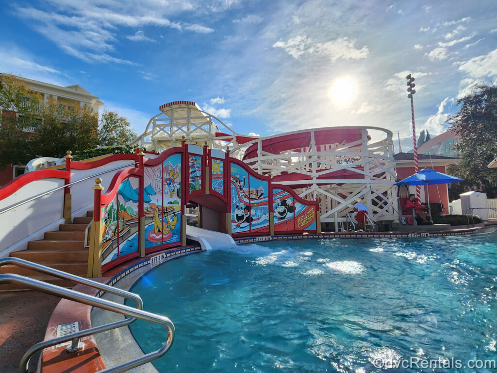 The Luna Park Pool at Disney’s BoardWalk Villas under sunny blue sky. The steps leading up to the Keister Coaster are visible under the colorful mural decorated with Mickey and Minnie Mouse, Pluto, Goofy, and Donald and Daisy Duck enjoying pool time.