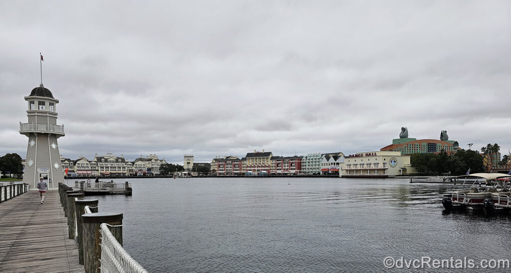 Disney’s BoardWalk Villas as seen from the wooden pier across Cresent Lake at Disney’s Beach Club Villas under a grey sky. On the right-hand side of the photo there are multiple burgundy and white boats in the marina.