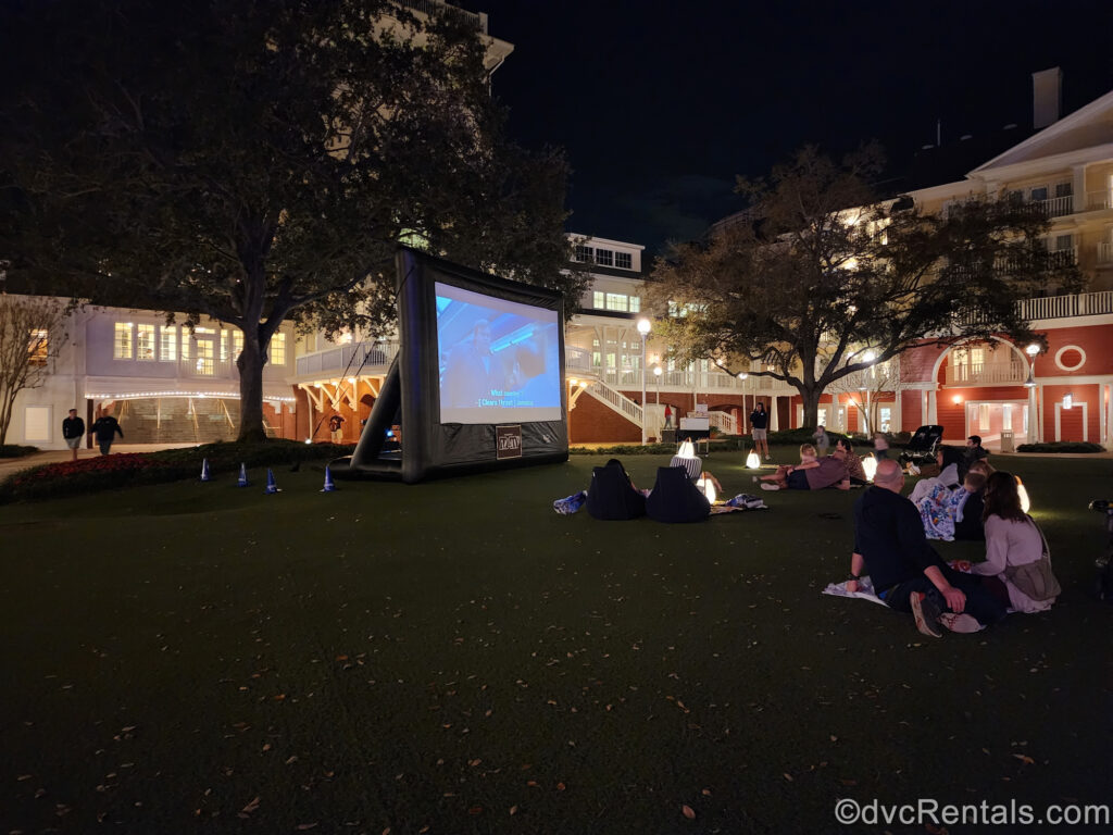 Small groups of guests sit on the grass to enjoy a Movie Under the Stars at Disney’s BoardWalk Villas at night. There is a large black inflatable screen showing a movie scene and the coral and white buildings of the resort and lit up in the background.