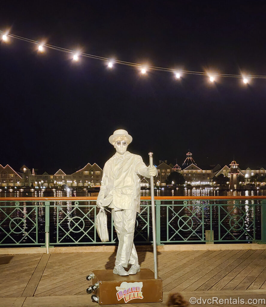 A performer dressed in a silver suit and hat with sunglasses and a white wig, meant to make them look like a statue, stands on the BoardWalk at night. Behind them you can see lights, Cresent Lake, and Disney’s Beach Club Villas across the water.