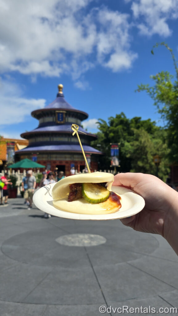 A hand holds a small white plate with a Crispy Duck Bao Bun on it, with the blue tiered building of the EPCOT China pavilion behind it. The bao bun is a soft white color with a green pickle, brown piece of duck, and hoisin sauce visible inside.