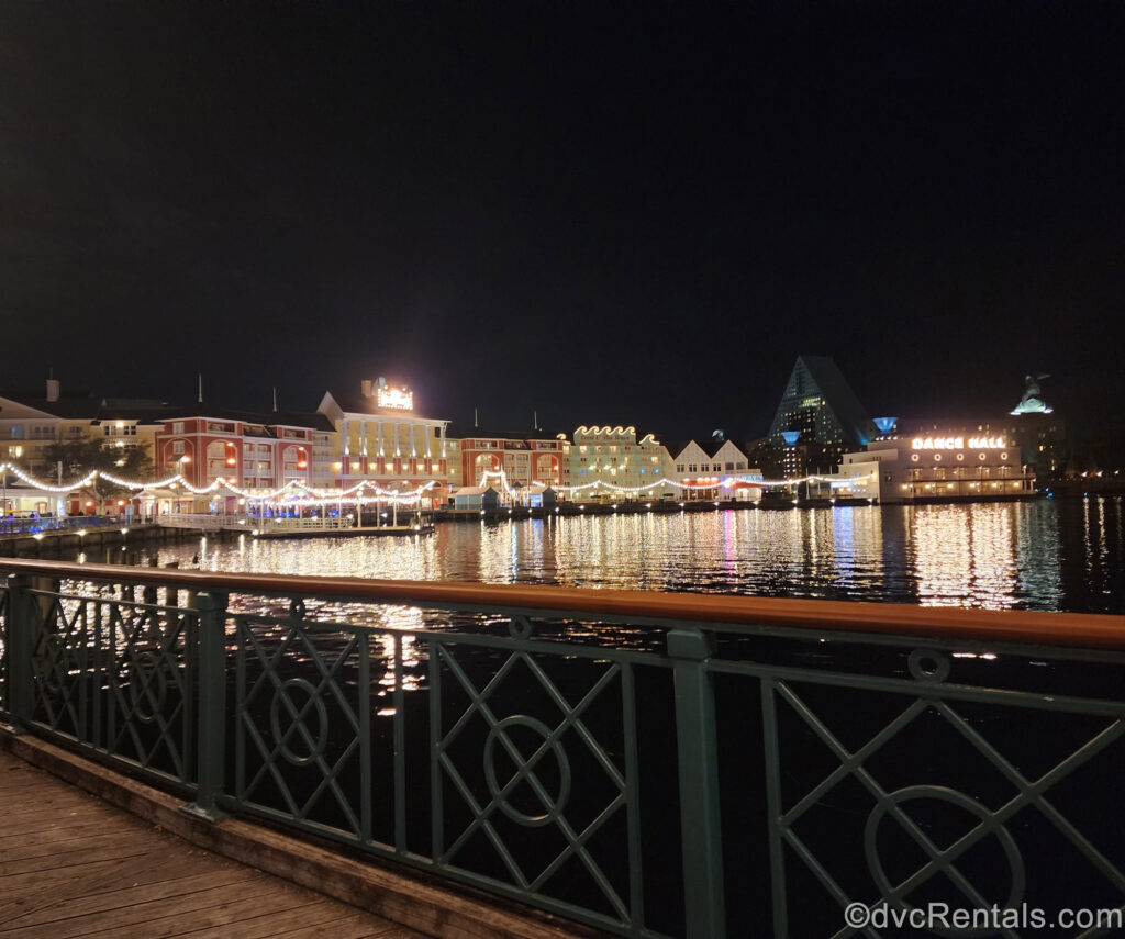 Disney’s BoardWalk Villas lit up at night. Small strands of lights glow along the row of the colorful resort buildings and reflect on Cresent Lake.