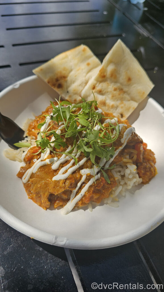 A small white plate on a black table holds a golden orange serving of Chicken Tikka Masala with fennel-spiced yogurt drizzled on it from the India booth. It is served over a bed of rice and two pieces of naan bread are behind it.