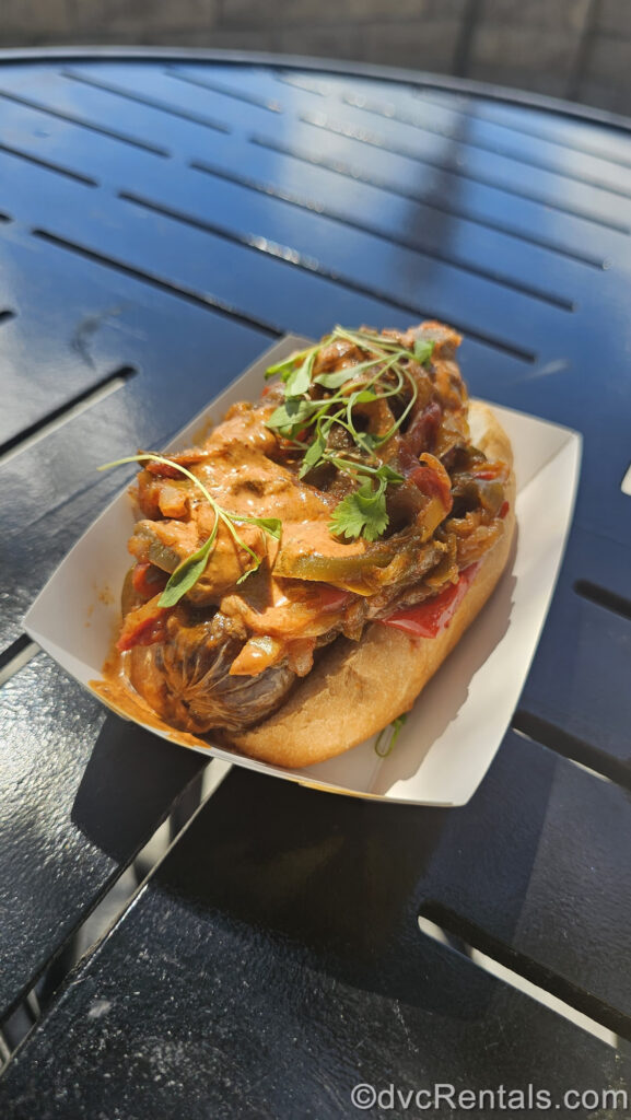 A small white basket on a black tabletop holds a serving of the Impossible Spicy Sausage. The sausage is served in a golden brown bun and is topped with Piri Piri peppers and onions and Piri Piri aioli.