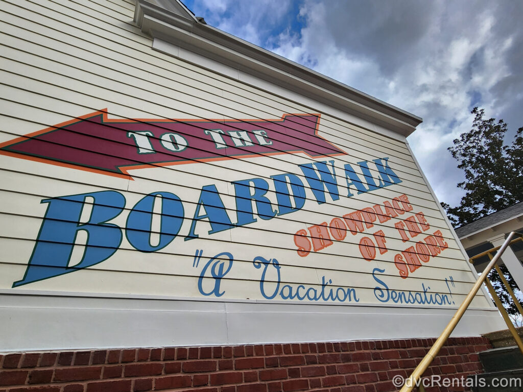 The phrases “To the BoardWalk, Showplace of the Shore” and “A Vacation Sensation!” are written in large blue and orange lettering on the pastel yellow side of one of the resort’s buildings. There is also a large, dark red arrow directing guests down towards the BoardWalk.