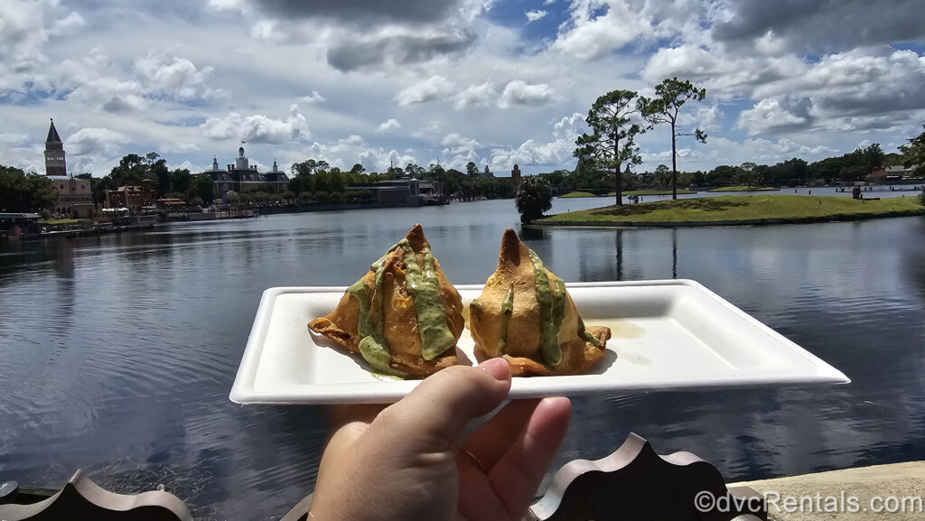 A hand holds a white plate with two Potato and Pea Samosas. The samosas are golden brown with a pea-green drizzle, and World Showcase Lagoon, landscaping, and buildings of various country pavilions are visible in the background.