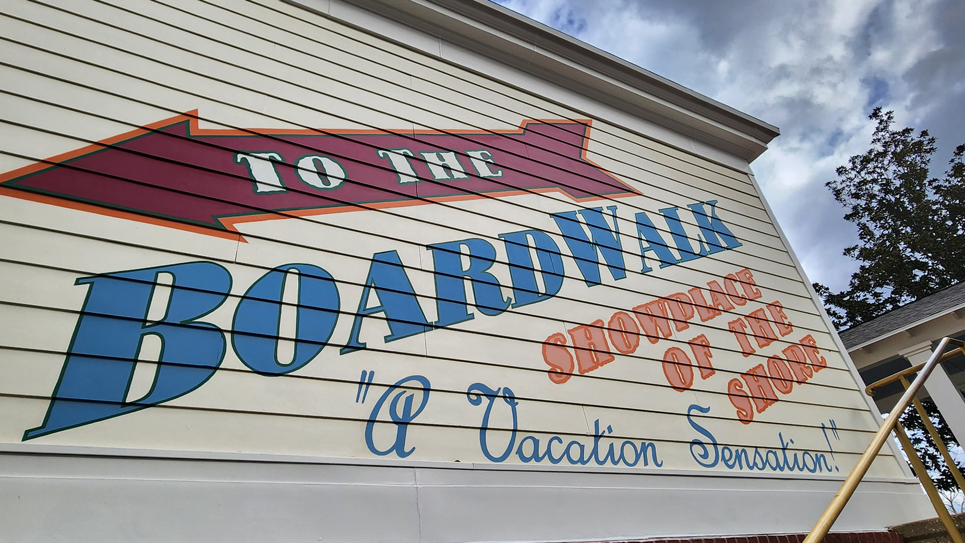 The phrases “To the BoardWalk, Showplace of the Shore” and “A Vacation Sensation!” are written in large blue and orange lettering on the pastel yellow side of one of the resort’s buildings. There is also a large, dark red arrow directing guests down towards the BoardWalk.