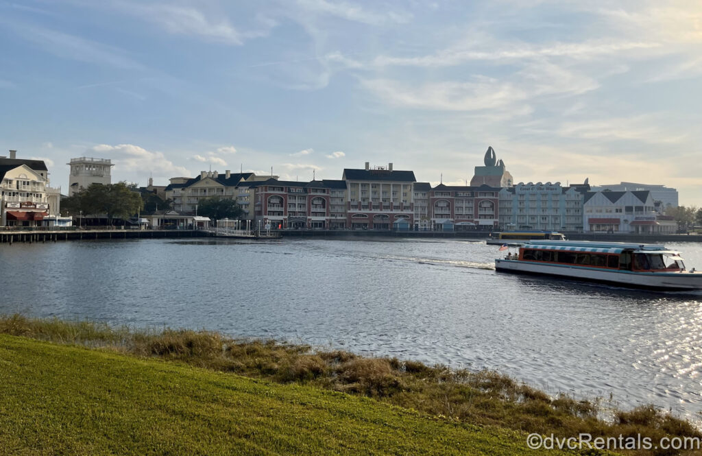 The colorful buildings of Disney’s BoardWalk Villas are seen at a distance as 2 boats drive by on the waters of Cresent Lake under a blue sky. The boat in the foreground is blue and white with pink accents, and the one further back is yellow and white.