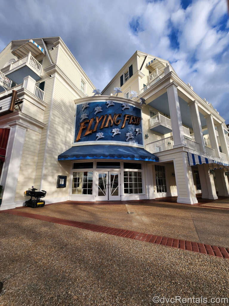 The blue exterior sign and awning of Flying Fish restaurant at Disney’s BoardWalk Villas between two light yellow resort buildings. The words “Flying Fish” are in orange with lots of fish with wings decorating the sign. Beneath the awing are the glass doors to the restaurant.