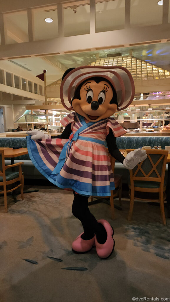 Minnie Mouse poses in her pink, purple, and blue beach-inspired dress and hat at Cape May Café at Disney’s Beach Club Villas. Behind her you can see blue and brown restaurant seating as well as some of the breakfast buffet area.