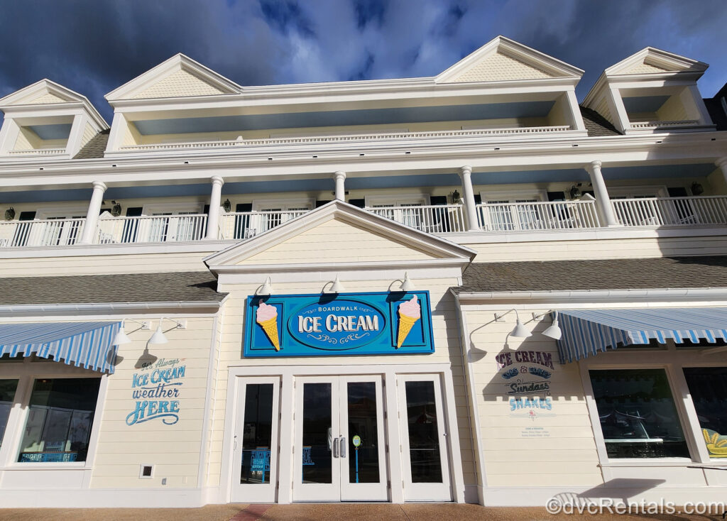 BoardWalk Ice Cream at Disney’s BoardWalk Villas. The building in light yellow with white trim, and there is a blue sign with the restaurant name on it and two pink ice cream cones. On the left side of the glass door entry there are the words “It’s always ice cream weather here” painted, and the words “Ice cream cups & cones, sundaes, shakes” are painted in blue on the right hand side.