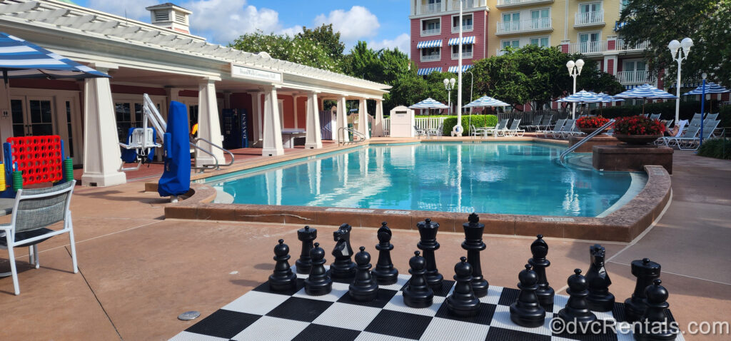 A life-sized black and white chess game is seen in front of the Community Hall area leisure pool at Disney’s BoardWalk Villas. The red and white Community Hall building is seen on the left, and there are several sun loungers and blue and white striped pool umbrellas on the right around the pool.