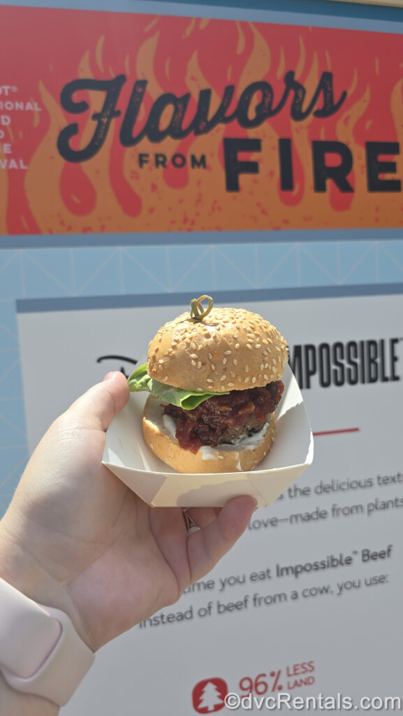 A hand holds a small white basket with the Impossible Montreal-Style Burger Slider from the Flavors From Fire booth. The slider is topped with cheddar cheese and tomato jam with a sesame seed bun.