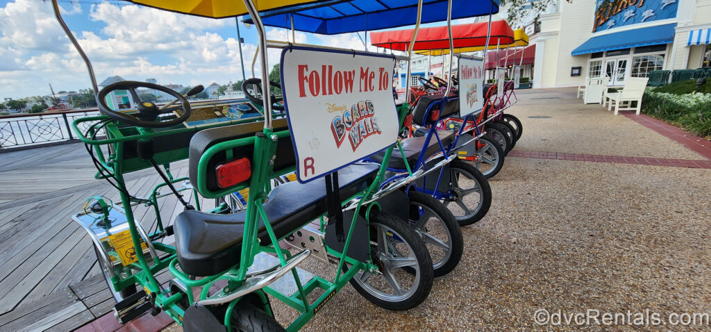 Green, blue, and red Surrey bikes in a line looking out to the BoardWalk. On the back of the bikes there are signs reading “Follow Me To Disney’s BoardWalk” in red and multicolored lettering.