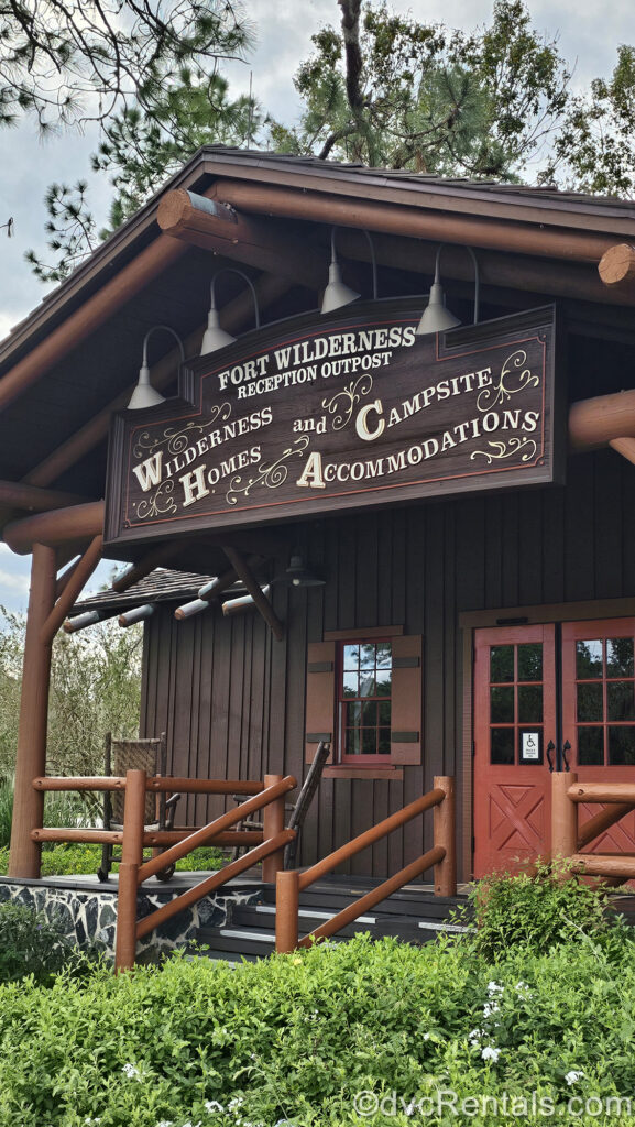 The dark brown log-cabin style exterior of the lobby at Disney’s Fort Wilderness Resort & Campground. Over the red doors there is a brown sign reading “Fort Wilderness Reception Outpost; Wilderness Homes and Campsite Accommodations in which script.