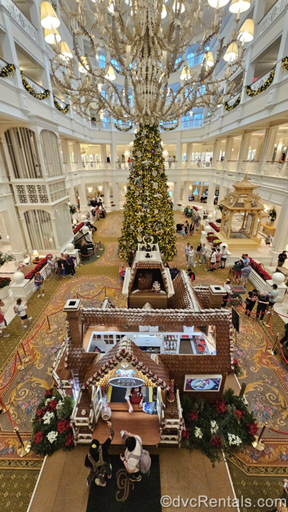 A life size gingerbread house and large Christmas tree with white lights and Victorian ornaments are displayed in the cream-colored main lobby at Disney’s Grand Floridian Resort & Spa, as seen from the second floor looking down.