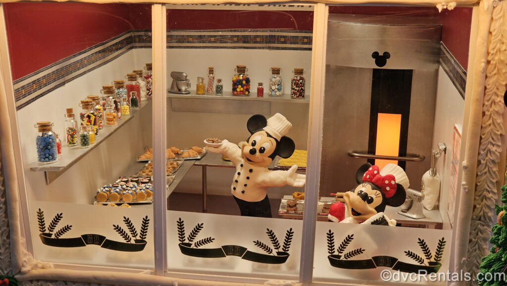 Small figures of Mickey and Minnie Mouse wearing chef hats are arranged to look as though they are baking cookies inside the gingerbread BoardWalk Deli replica on display at Disney’s BoardWalk Villas.