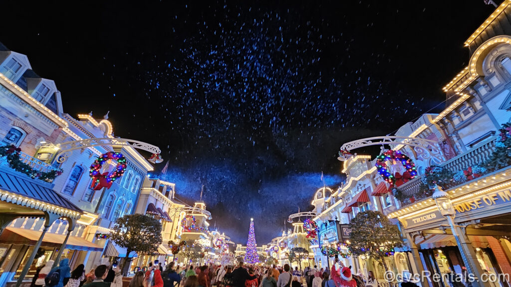 Garlands, wreaths, and a large Christmas tree with multicolored lights and ornaments decorate the buildings of Main Street USA at night as white pretend snow falls over the crowd of guests.