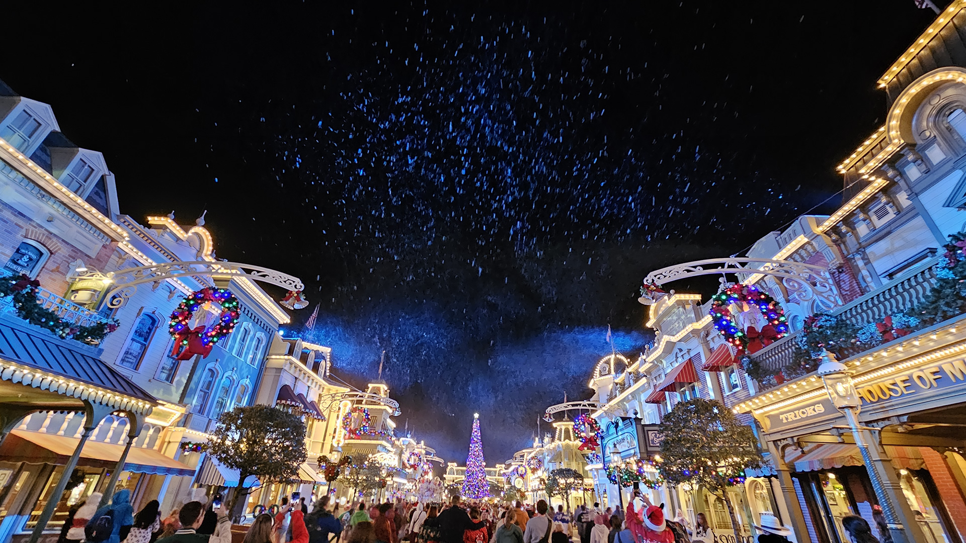 Garlands, wreaths, and a large Christmas tree with multicolored lights and ornaments decorate the buildings of Main Street USA at night as white pretend snow falls over the crowd of guests.