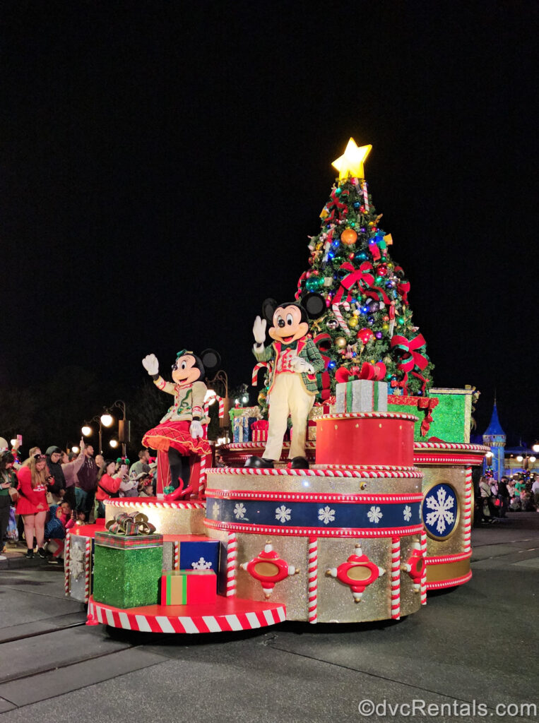 Mickey Mouse and Minnie Mouse perform during Mickey’s One Upon a Christmas Parade at Magic Kingdom. The characters wear beige, green, and red holiday outfits, and the float is decorated with a multicolored Christmas tree and giant gift boxes.