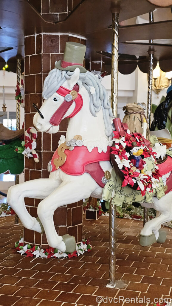 A White carousel horse made of gingerbread is meant to resemble Ebenezer Scrooge from Mickey’s Christmas Carol. The horse has a red saddle, green top hat, black-rimmed glasses, and a red and white Christmas wreath decorated with pretend dollar bills and a figurine of Scrooge McDuck as the character from the film.