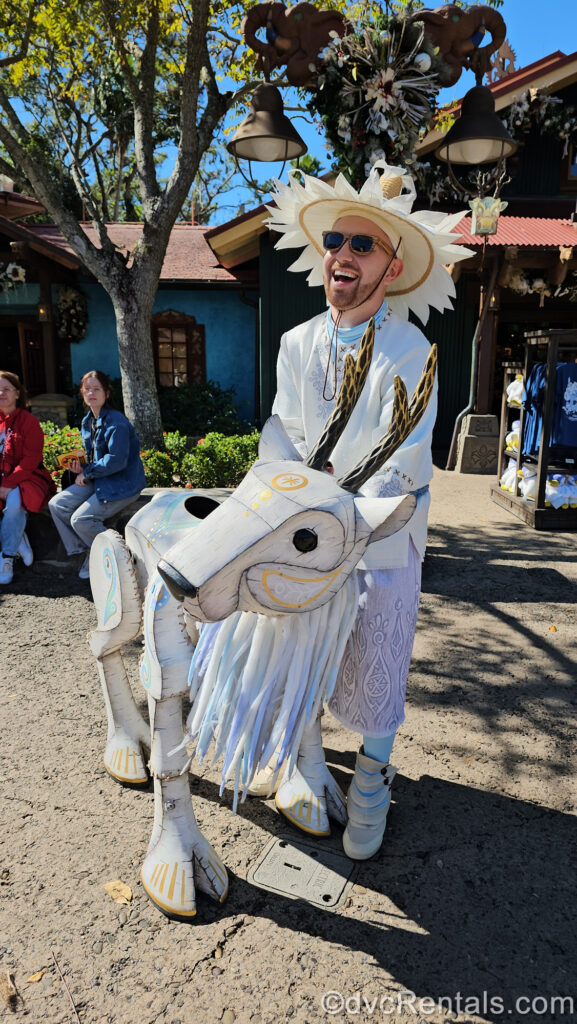 A happy puppeteer in a white, wintery costume plays with a white mountain goat puppet. The puppet is decorated with light blue and gold designs.