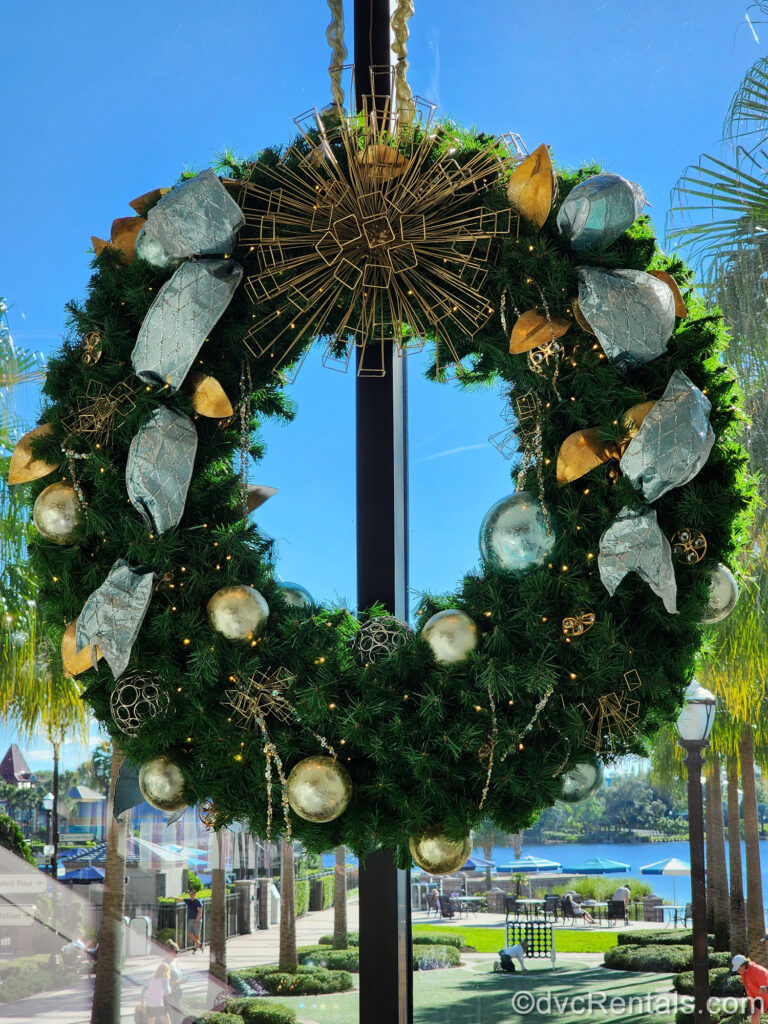 A large green Christmas wreath decorated with gold, pewter, and light blue ornaments and ribbons hangs in front of a window at Disney’s Riviera Resort. The resort grounds and water can be seen in the background.