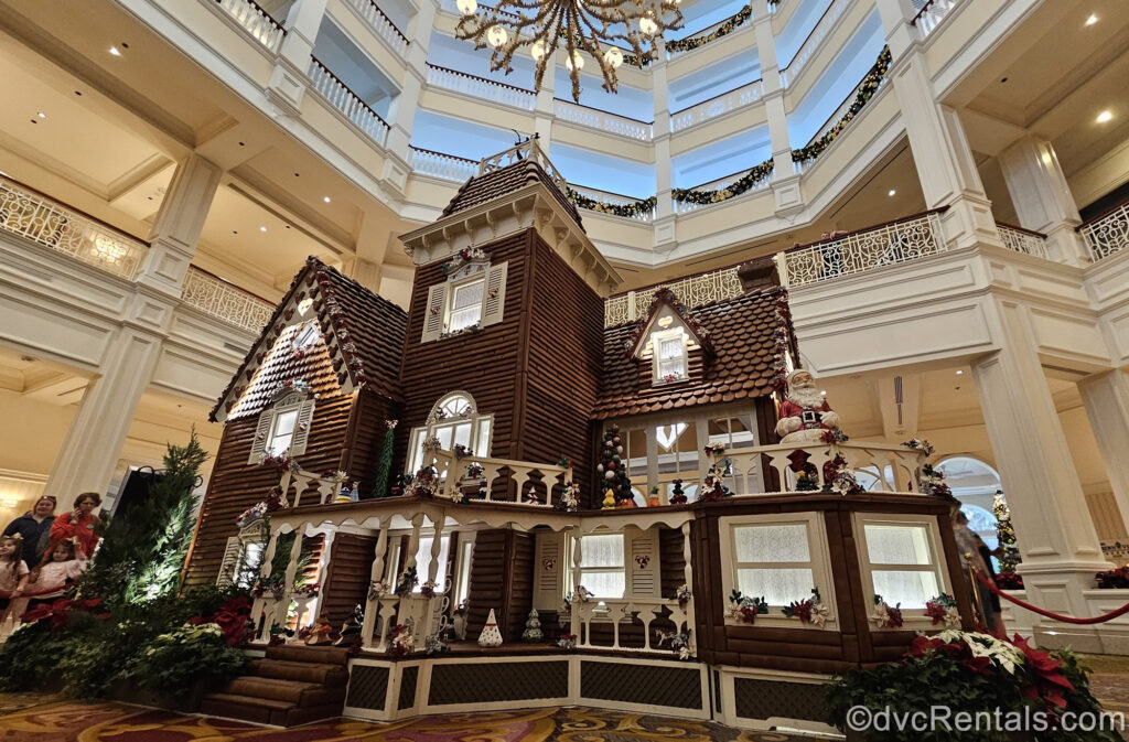 A life-size gingerbread house with white trim, decorated with multicolored miniature Christmas trees and a Santa statue is displayed at Disney’s Grand Floridian Resort & Spa.