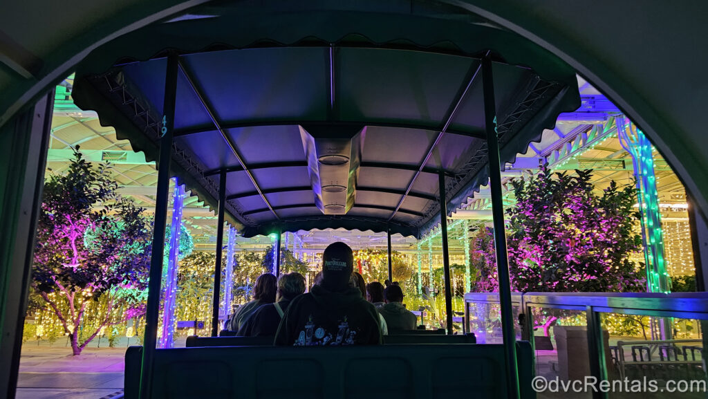 A green Living with the Land boat carrying guests enters a greenhouse where the plants and shelving are decorated with white, purple, green, blue, and pink twinkling Christmas lights.