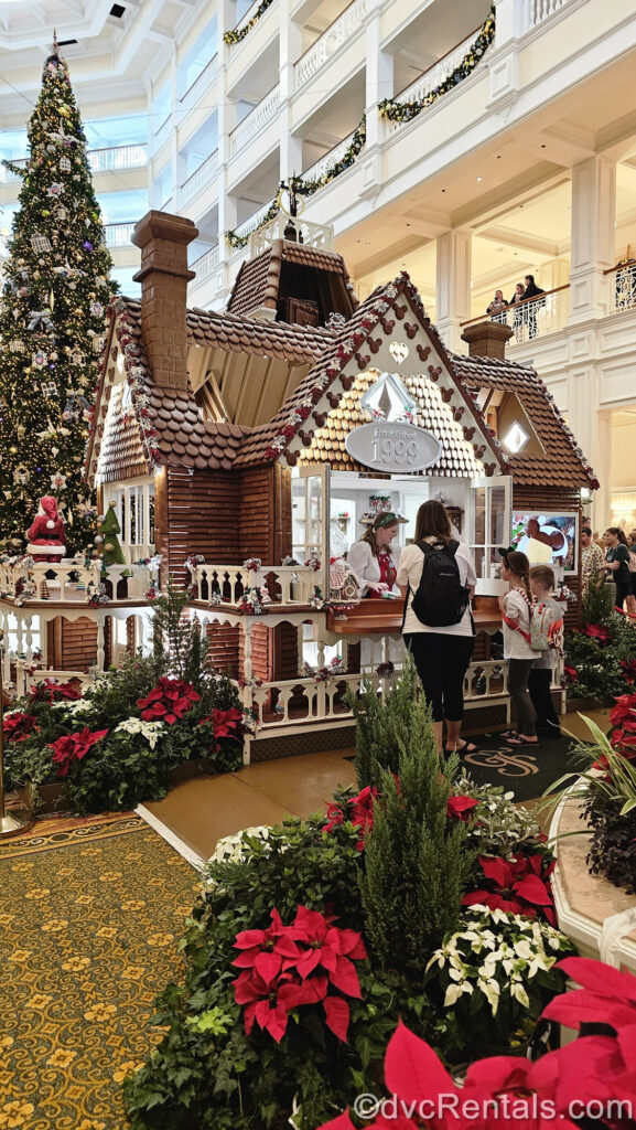 Guests are seen purchasing holiday treats at the window of the life-size gingerbread house at Disney’s Grand Floridian Resort & Spa. Behind the gingerbread house is a giant Christmas tree with pastel ornaments, and the gingerbread house is also surrounded by red, green, and white poinsettias.