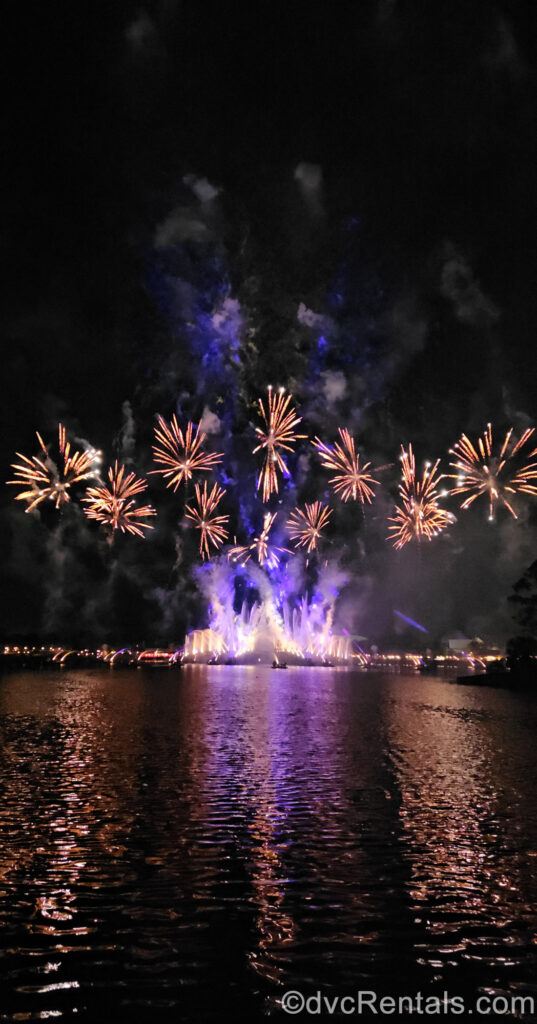 Golden fireworks erupt in the sky over the white, purple, and pink lights and fountains of the Luminous The Symphony of Us nighttime show in World Showcase Lagoon at EPCOT.