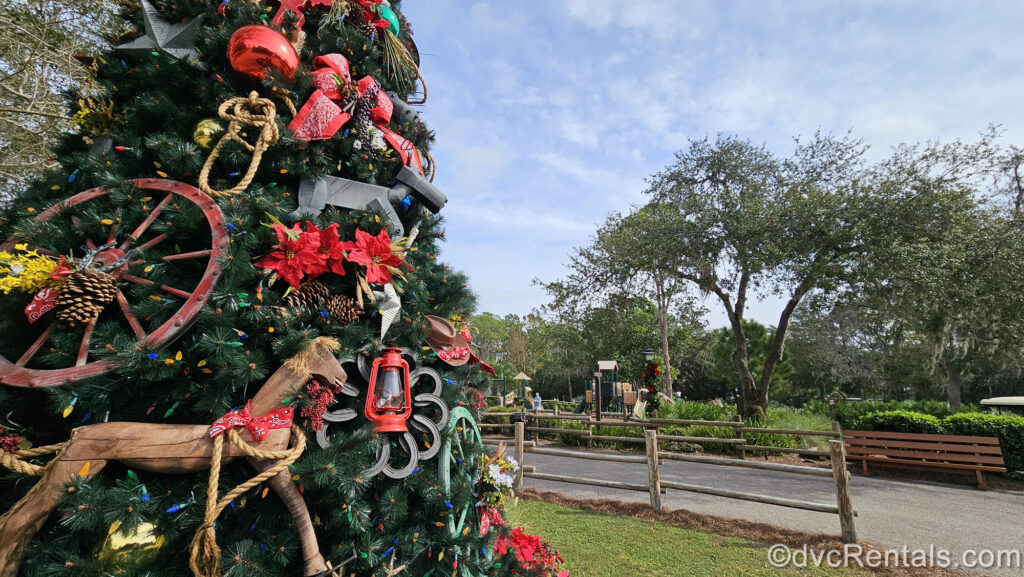 A green Christmas tree with frontier-style ornaments like a wooden horse and wagon wheel is seen outside under sunny skies at Disney’s Fort Wilderness Resort & Campground. To the right of the tree, resort grounds with a wooden bench and playground can be seen in the distance.