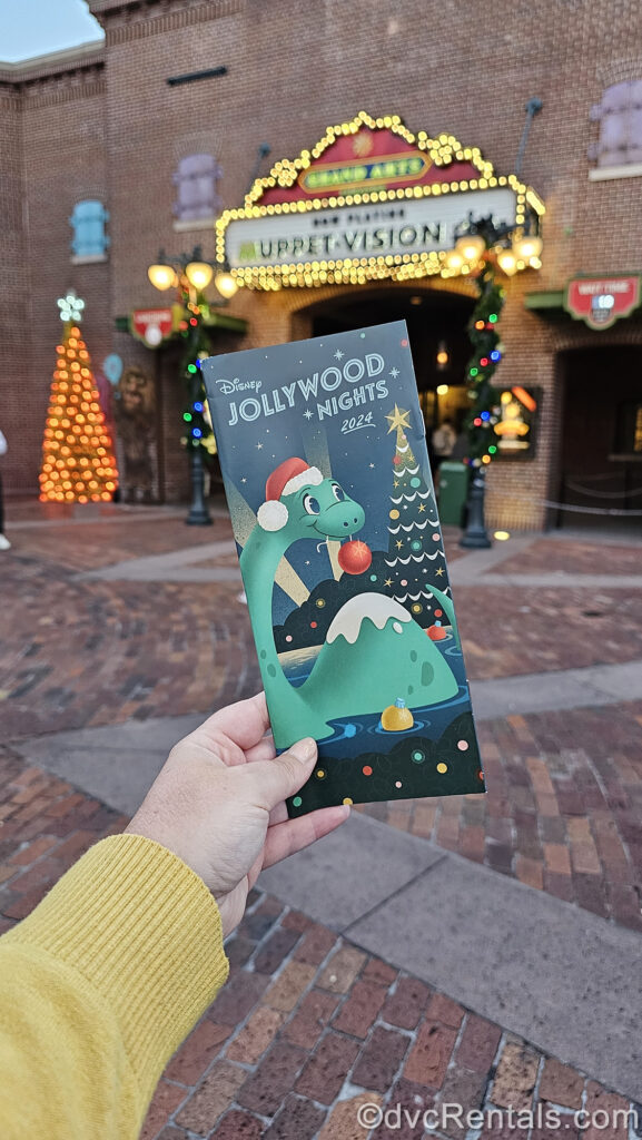 A hand holds the green Disney Jollywood Nights 2024 map featuring a cartoon version of Gertie the dinosaur in a red Santa hat in front of a Christmas tree. The map is held up in front of the lit-up marquee for Muppet*Vision 3D and colorful Christmas decorations.