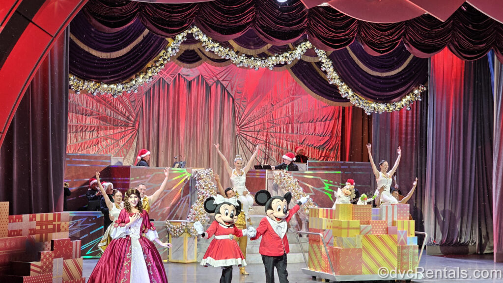 Belle, Minnie, Mickey, Miss Piggy, Kermit the Frog, and an ensemble of dancers and musicians in holiday outfits perform on a stage decorated with red and gold gift boxes and silver and gold ornaments during the Disney Holidays in Hollywood show.