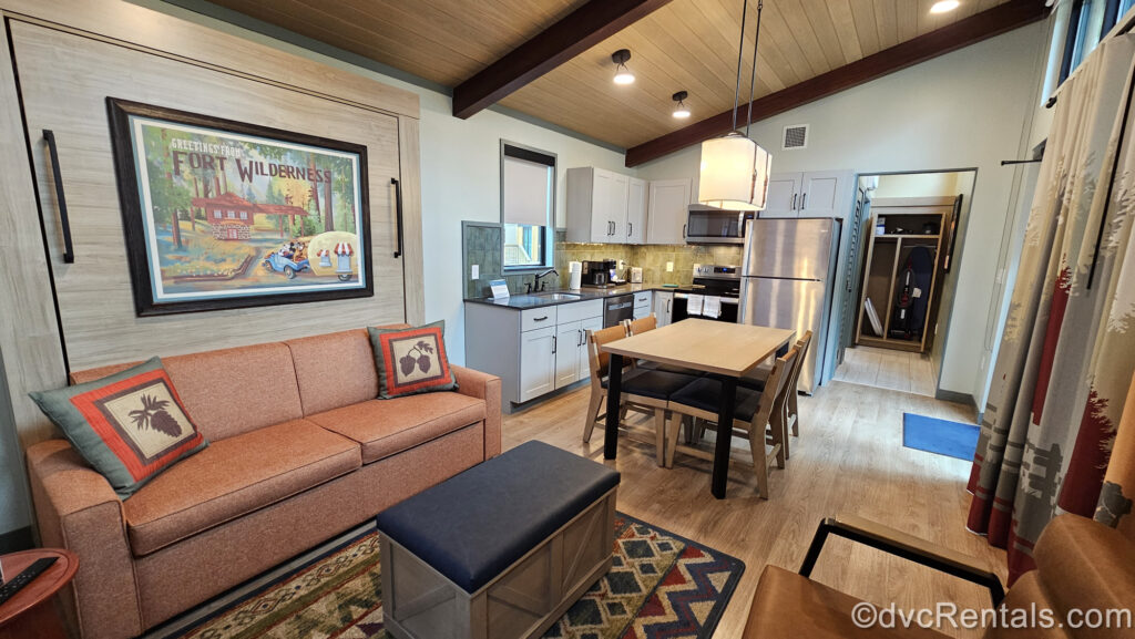 Interior of a One Bedroom Cabin. The walls are cream colored with light wood paneling on the ceiling and behind the orange sofa. There is a Fort Wilderness mural with Mickey driving a camper above the couch, a full kitchen with white cabinets, and wooden dining table.