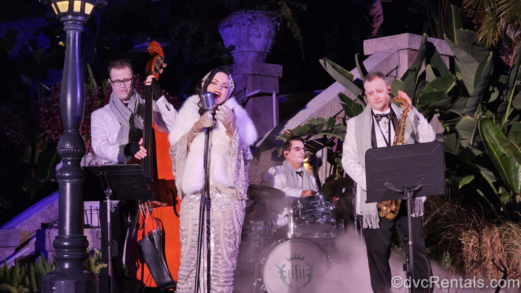 A 4-person group plays music outside Tower of Terror at Hollywood Studios. The female lead singer is dressed in a sequined white and silver gown and white fur shawl, and the 3 men playing instruments in the back wear white and black suits as fog rolls over the stage.